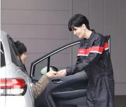 黒と赤のユニフォームを着用した整備士が、車両のドアを開けて顧客と対応している様子。車両の受け渡し場面
