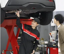 整備工場にて、リフトアップされた車両の下で点検作業を行う整備士。黒と赤のユニフォームを着用し、顧客も作業の様子を確認している。赤いリフトが特徴的