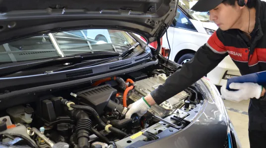整備士がエンジンルームの点検作業を行う様子。黒と赤のNISSANユニフォームを着用した整備士が、開けられたボンネットの中を確認している