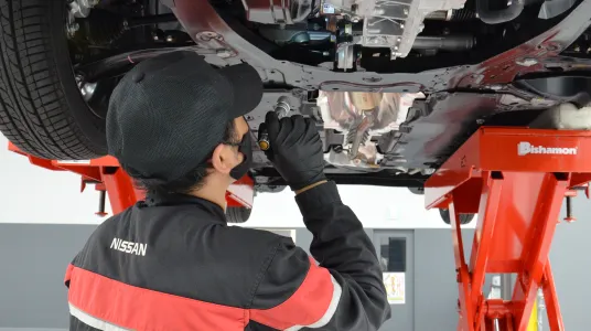 リフトアップされた車両の下で点検を行う整備士。黒と赤のNISSANユニフォームと黒い手袋を着用し、車両下部のアンダーカバーを確認している。赤いリフトが特徴的