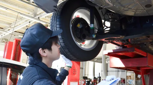 整備士がリフトアップされた車両のタイヤ周りを点検している様子。ネイビーの作業着とキャップを着用した整備士が、ライトを使用して足回りを入念に確認している