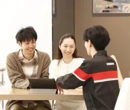 商談スペースでの接客シーン。黒と赤のストライプ入りユニフォームを着た整備士が、カジュアルな服装のカップルに対してタブレットを使用しながら説明を行っている様子。温かみのある照明の室内で、和やかな雰囲気の商談風景