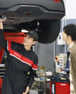 自動車整備士がリフトアップされた車両の下で車検点検作業を行う様子。整備士は黒と赤のツートンカラーのユニフォームを着用
